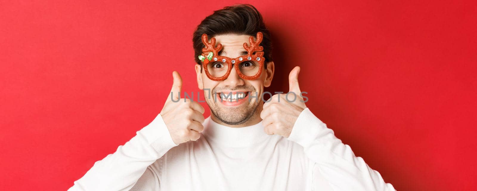 Concept of winter holidays, christmas and celebration. Close-up of excited handsome man in party glasses, smiling and showing thumbs-up in approval, standing over red background by Benzoix