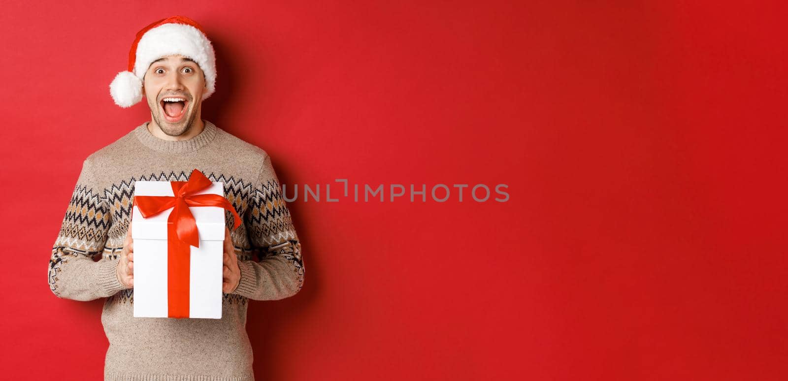 Image of excited handsome man receiving christmas gift, wearing santa hat and winter sweater, shouting for joy, holding present and standing over red background.