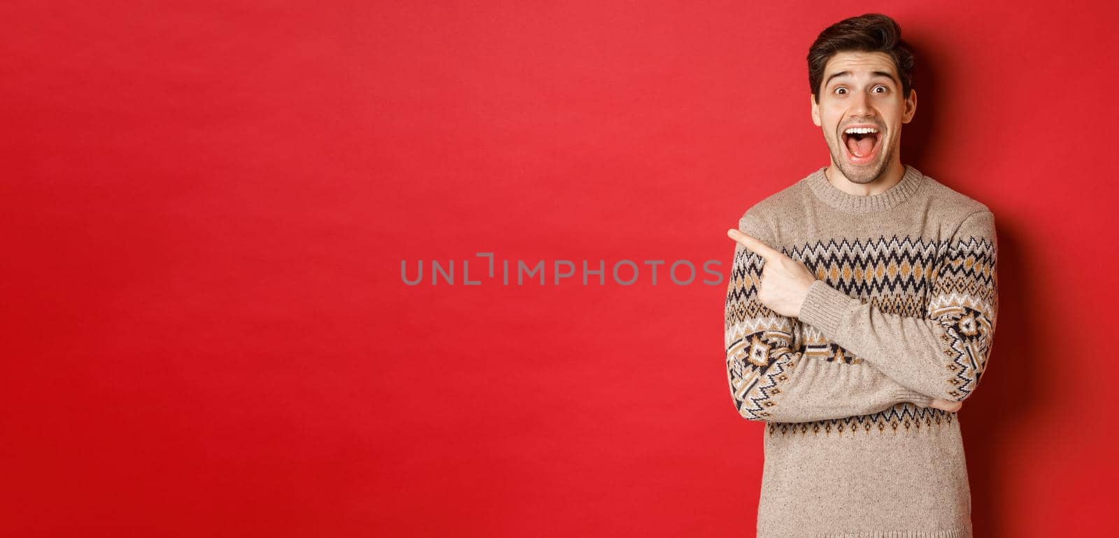 Portrait of amazed handsome guy, wearing christmas sweater, pointing finger at upper left corner logo and looking excited, showing awesome new year promo offer, standing over red background.