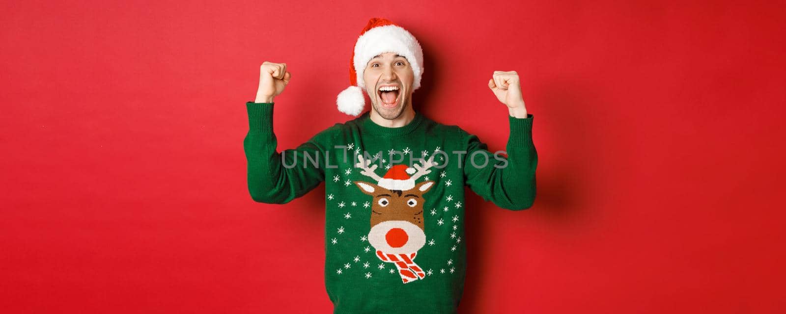 Portrait of cheerful attractive man celebrating new year, wearing green sweater and santa hat, shouting for joy, winning or triumphing, standing over red background.