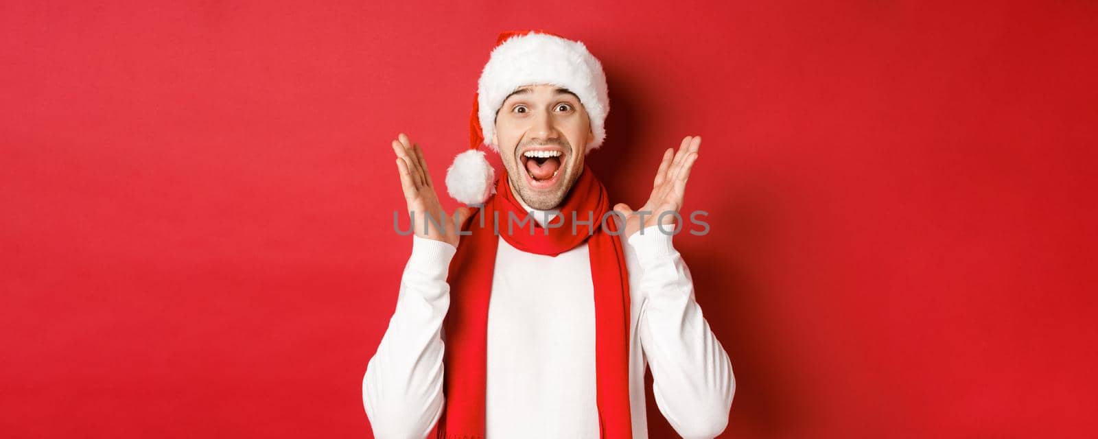 Concept of christmas, winter holidays and celebration. Image of handsome man looking surprised at new year promo offer, smiling amazed, standing over red background by Benzoix