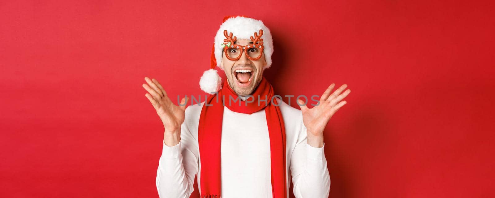Concept of christmas, winter holidays and celebration. Image of surprised and happy man looking amazed, wearing party glasses and enjoying new year, standing over red background by Benzoix
