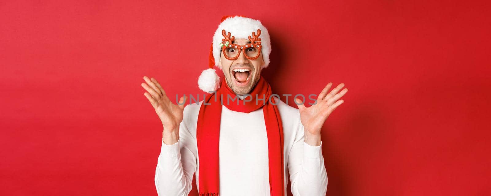 Concept of christmas, winter holidays and celebration. Image of surprised and happy man looking amazed, wearing party glasses and enjoying new year, standing over red background.