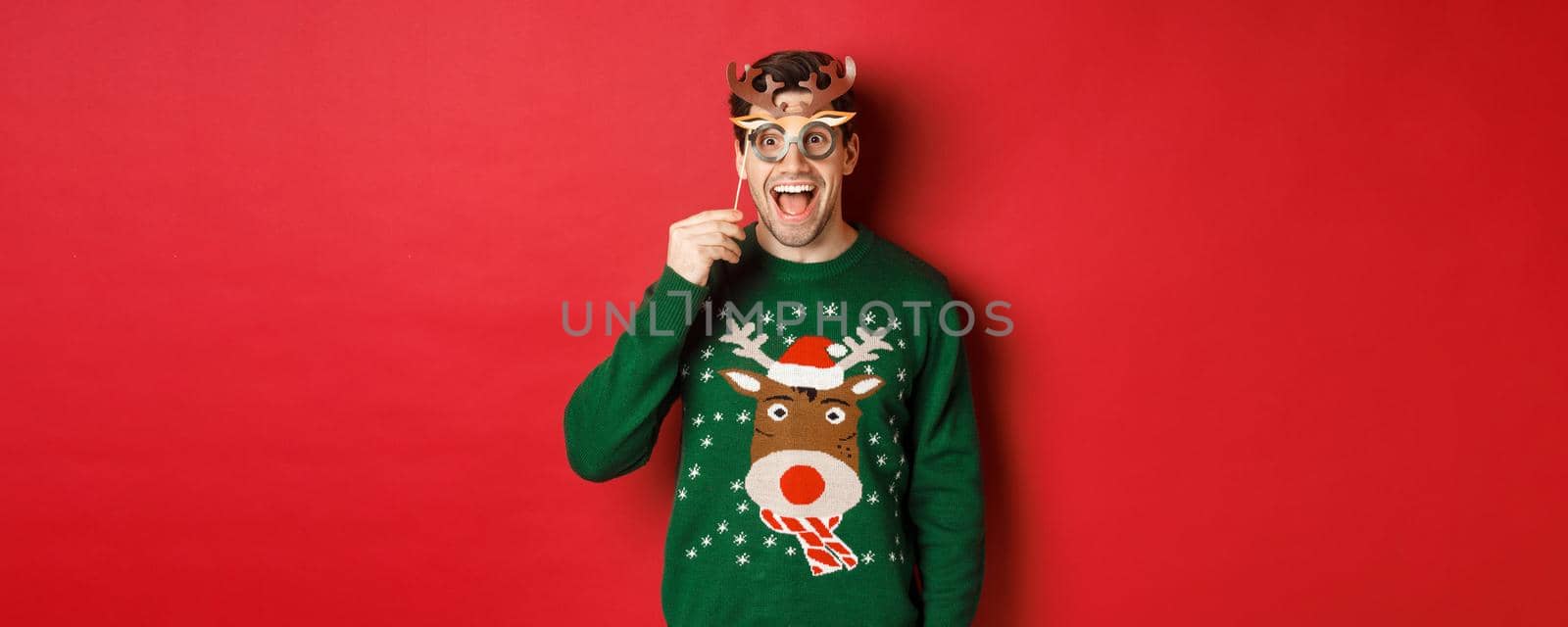 Handsome surprised man in christmas sweater, holding party mask and smiling, enjoying new year celebration, standing over red background by Benzoix