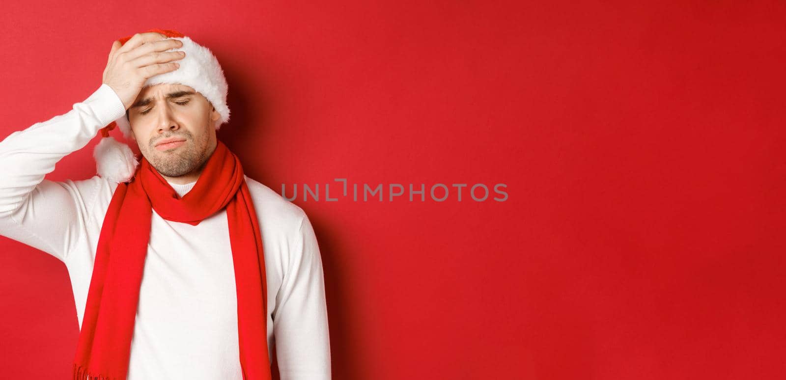 Concept of christmas, winter holidays and celebration. Portrait of sad and frustrated man in santa hat, feeling bad, standing over red background.