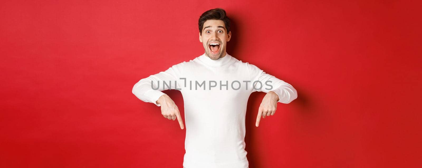 Image of amazed handsome guy in white sweater, reacting excited to christmas advertisement, pointing fingers down, showing logo, standing over red background by Benzoix