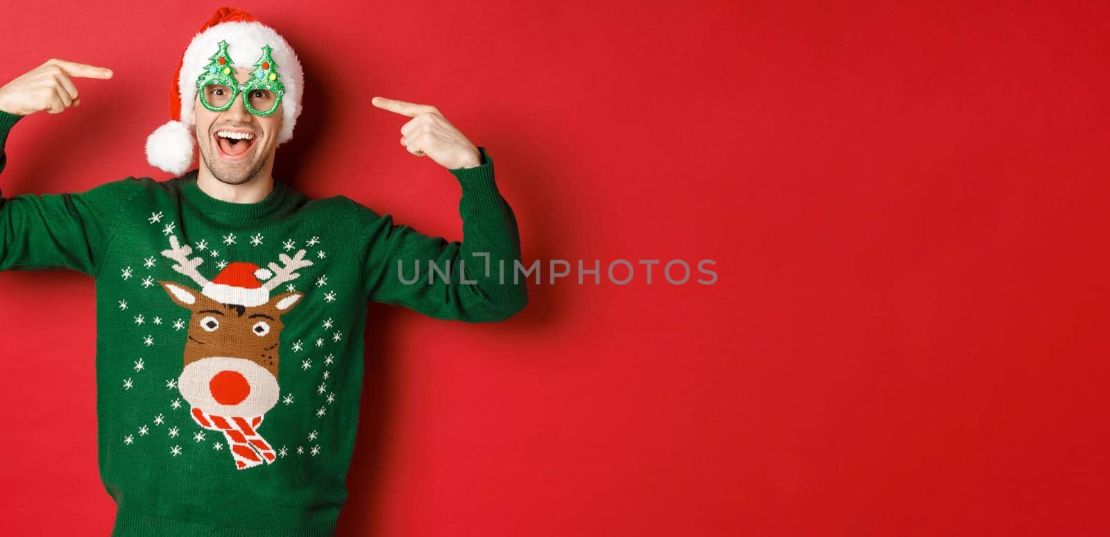 Image of excited smiling guy pointing at party glasses and celebrating new year, standing joyful in green sweater and santa hat against red background.
