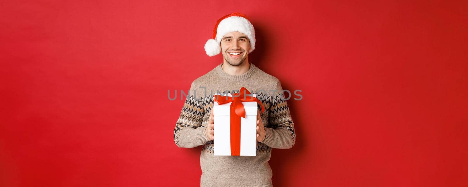 Image of handsome smiling man in santa hat and winter sweater, holding a present, giving christmas gift and wishing happy holidays, standing over red background.