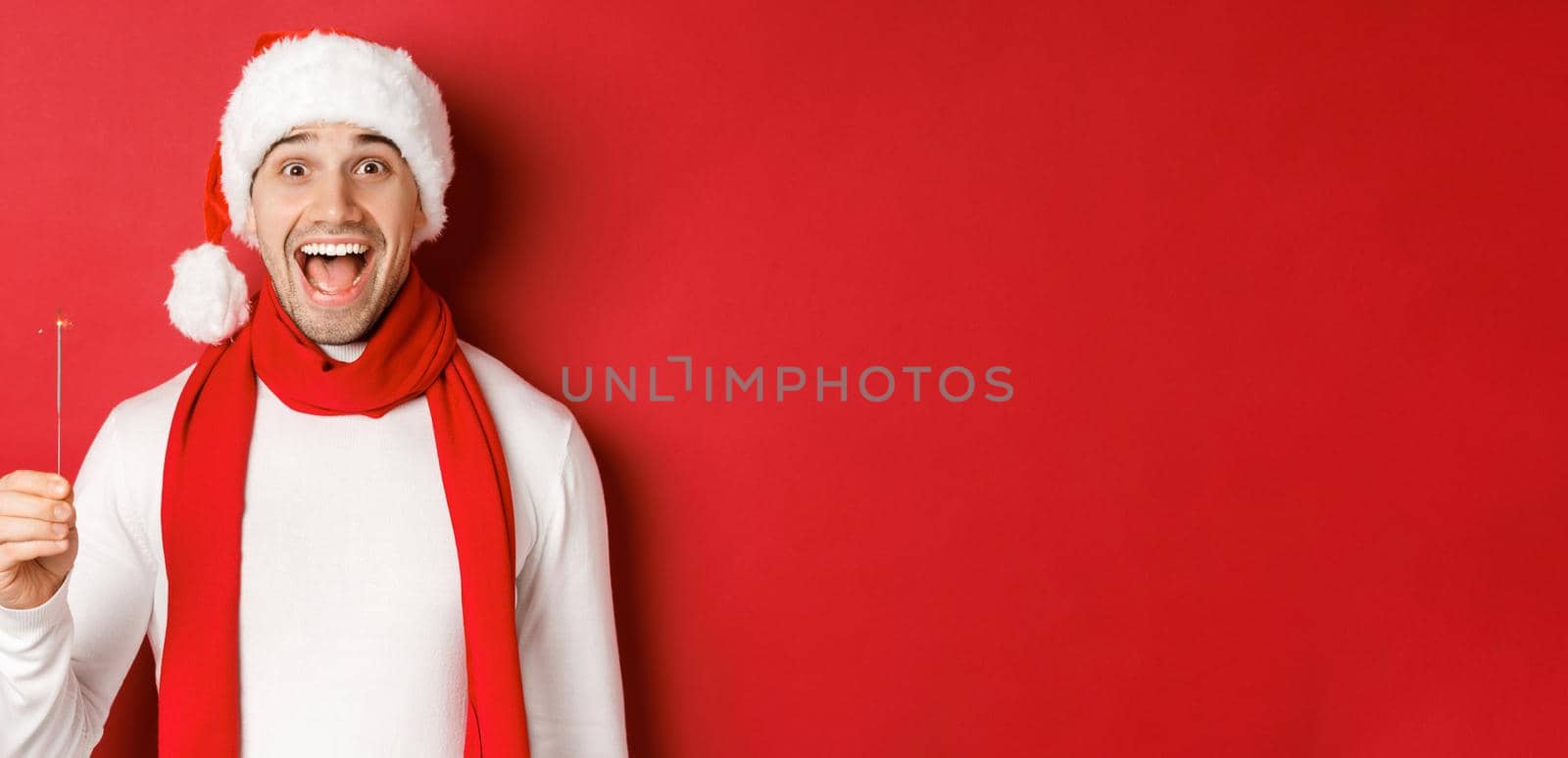 Concept of christmas, winter holidays and celebration. Close-up of happy handsome man in santa hat and scarf, having fun on new year party, holding sparkler and smiling, red background by Benzoix