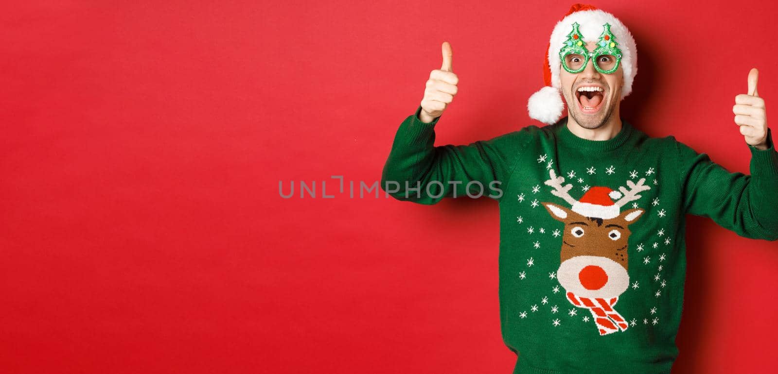 Portrait of super happy young man in party glasses, santa hat and sweater, showing thumbs-up in approval, recommending new year promo offer, standing over red background by Benzoix