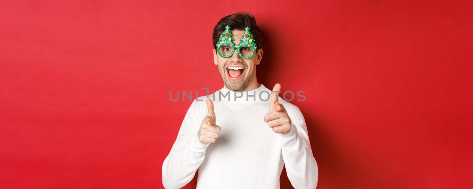 Joyful caucasian guy in party glasses and white sweater, smiling and pointing fingers at camera, wishing merry christmas and happy new year, standing over red background.