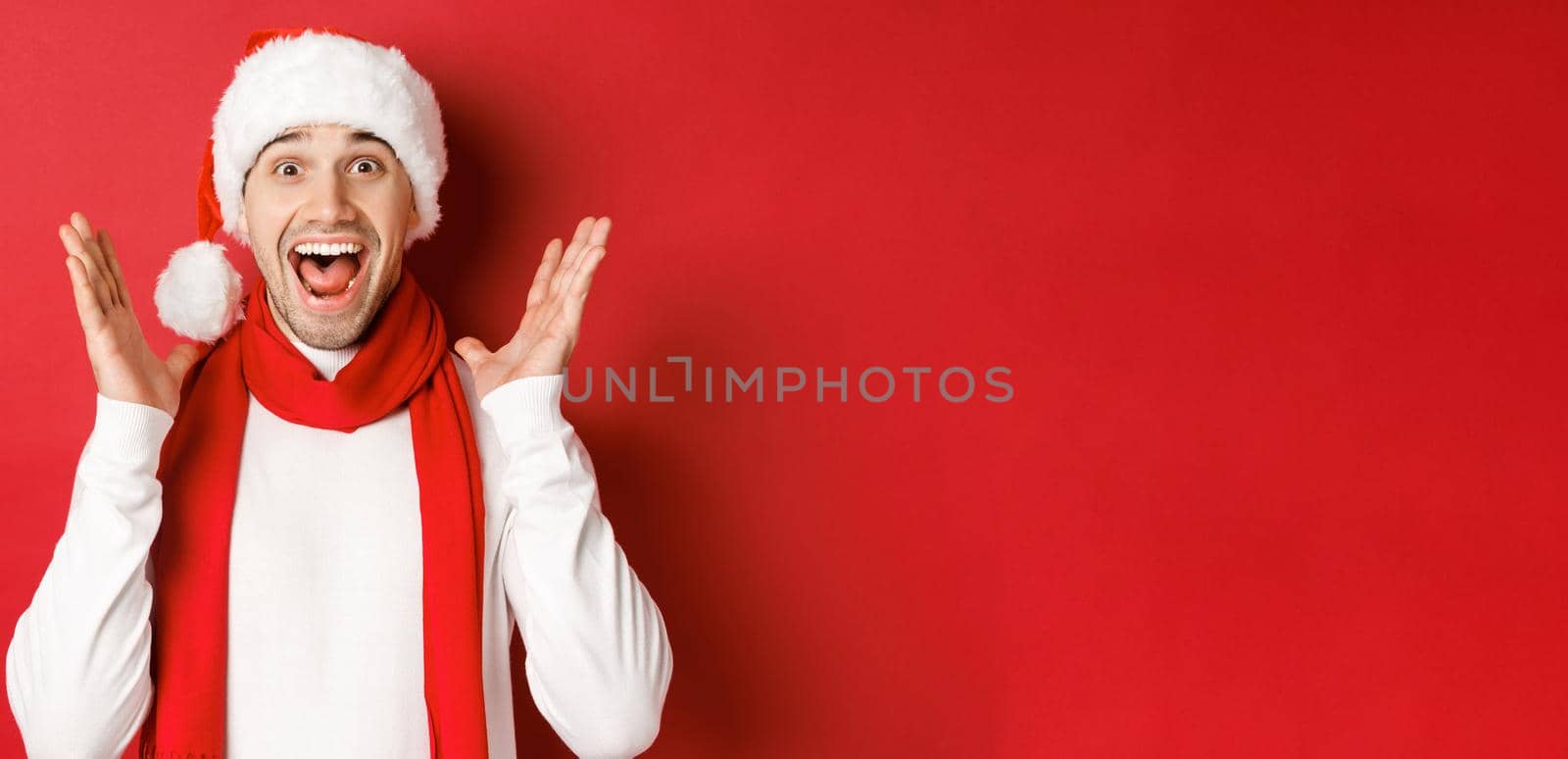 Concept of christmas, winter holidays and celebration. Image of handsome man looking surprised at new year promo offer, smiling amazed, standing over red background.