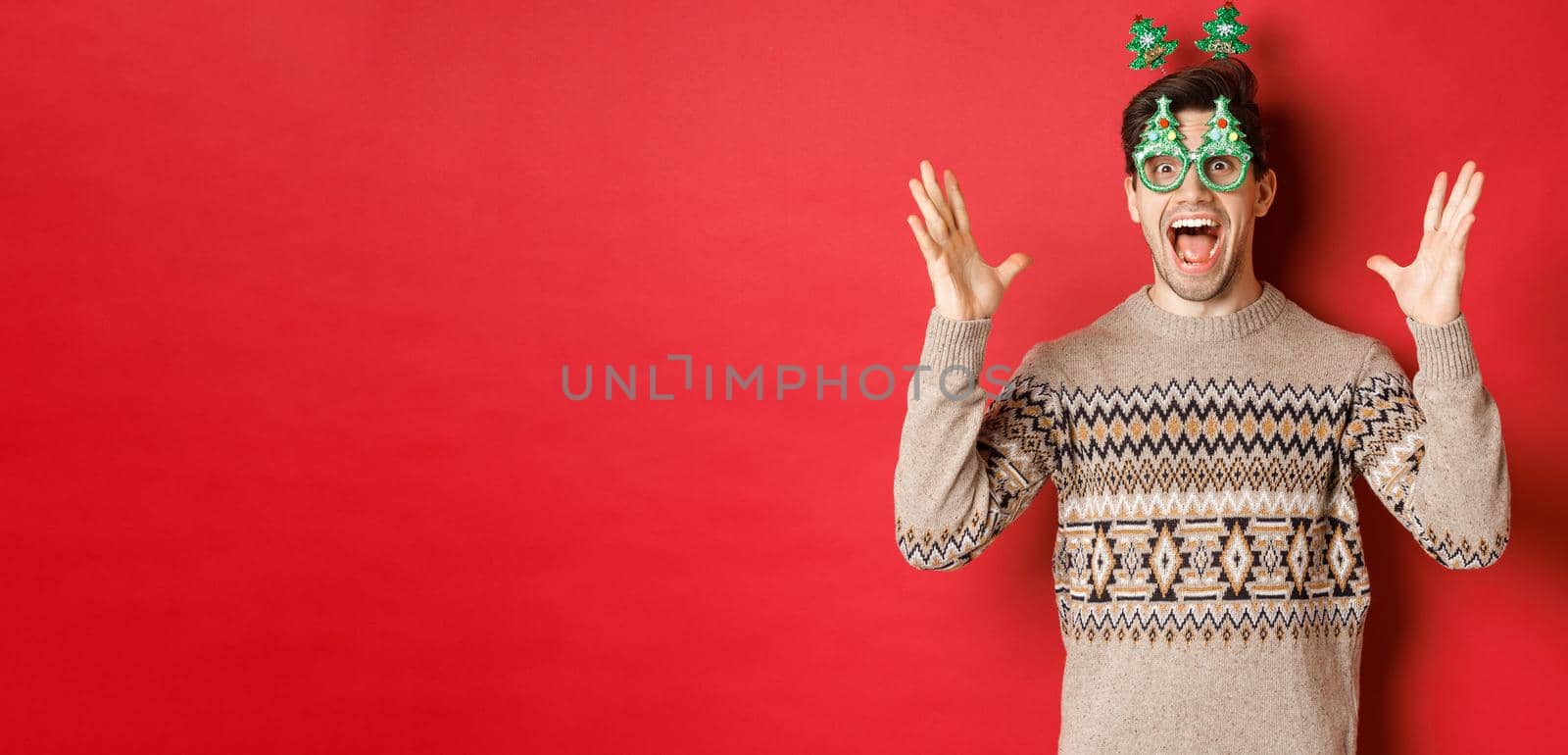 Image of excited and amazed caucasian guy in party glasses, christmas sweater, raising hands up and making big announcement, enjoying new year celebration, red background.