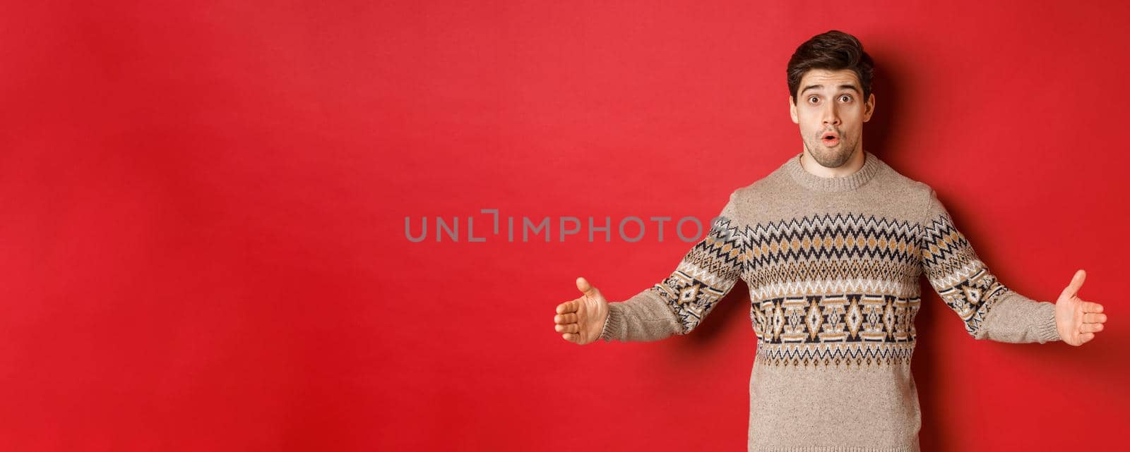 Image of amazed handsome man showing size of christmas present, spread hands sideways and shaping something large, standing over red background.