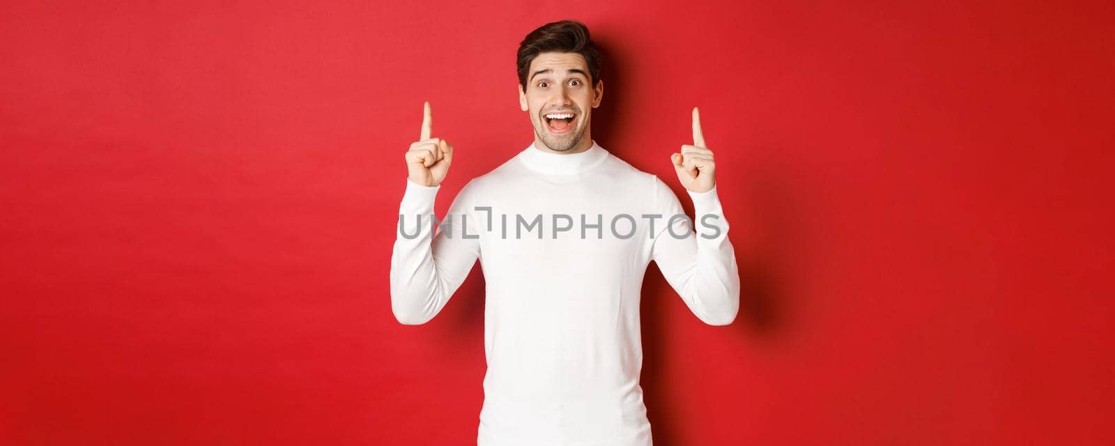 Concept of winter holidays. Good-looking young man with bristle, wearing white sweater, showing christmas advertisement on copy space, pointing fingers up and looking amazed, red background by Benzoix