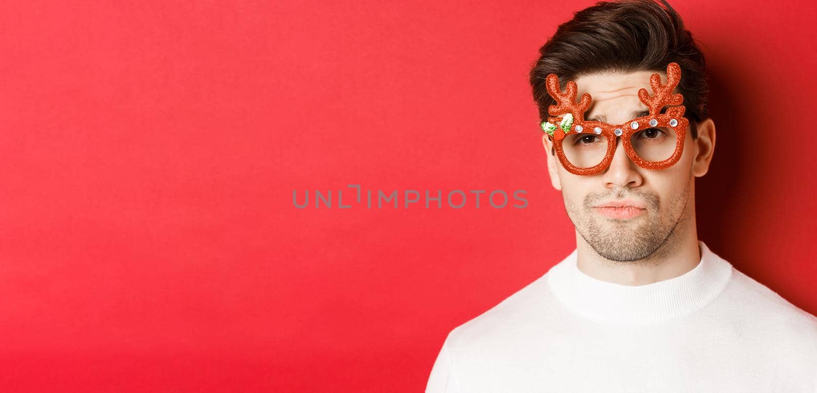 Concept of winter holidays, christmas and celebration. Close-up of skeptical handsome guy, wearing party glasses, smirk and looking unamused, standing over red background.