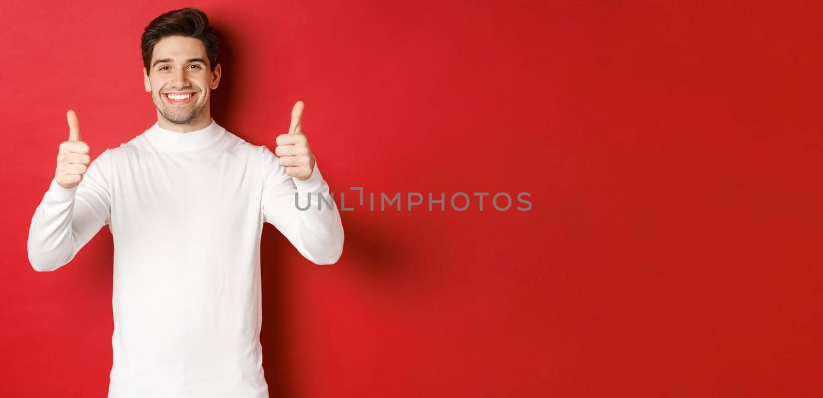 Concept of winter holidays, christmas and lifestyle. Portrait of satisfied handsome man in white sweater showing thumbs-up, praise something good, like it, standing over red background.