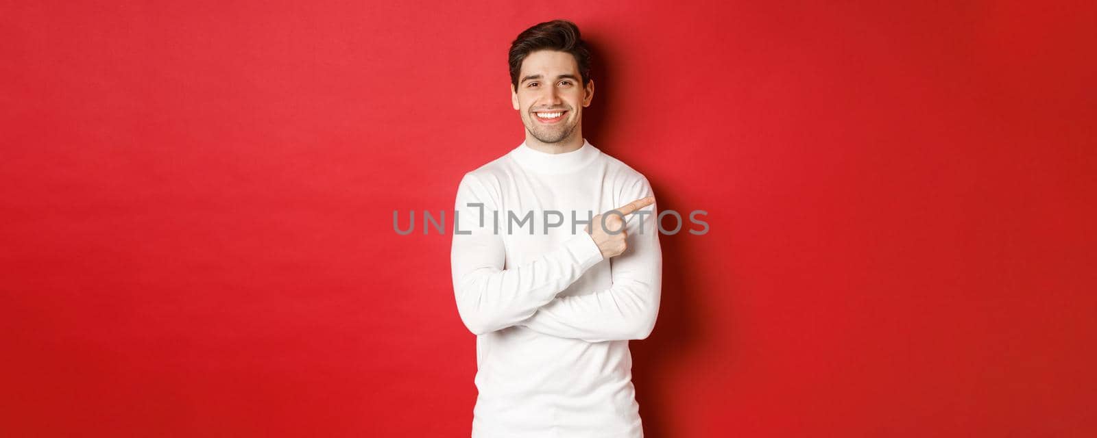 Handsome young man with bristle, wearing white sweater, pointing fingers at upper right corner and smiling, showing christmas banner, standing over red background.