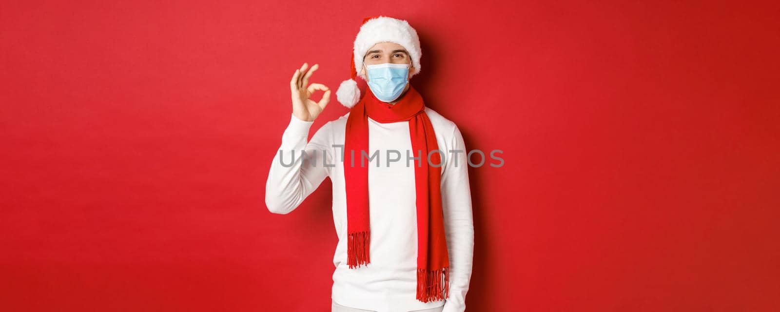 Concept of covid-19, christmas and holidays during pandemic. Portrait of happy and satisfied man in medical mask and santa hat, showing okay sign in approval, recommending something by Benzoix