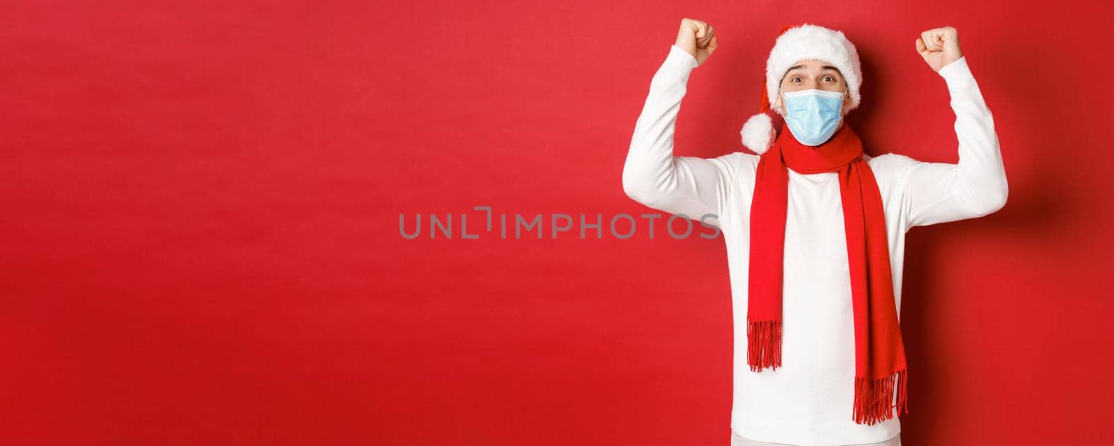 Concept of covid-19, christmas and holidays during pandemic. Portrait of happy man in santa hat and medical mask, rejoicing and celebrating new year, standing over red background.