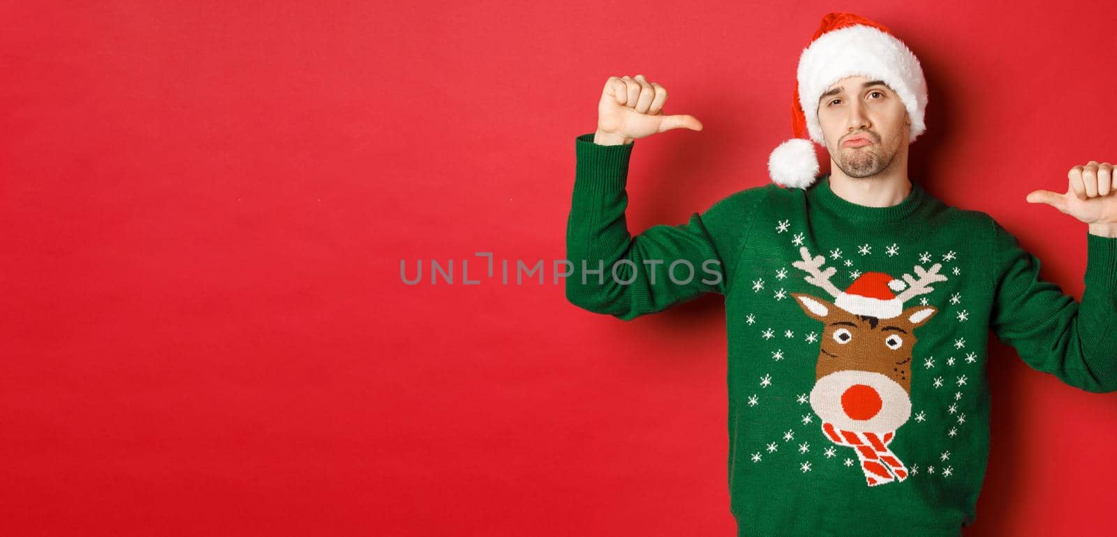 Image of handsome and confident young man in green sweater and santa hat, pointing at himself, celebrating christmas, standing over red background.