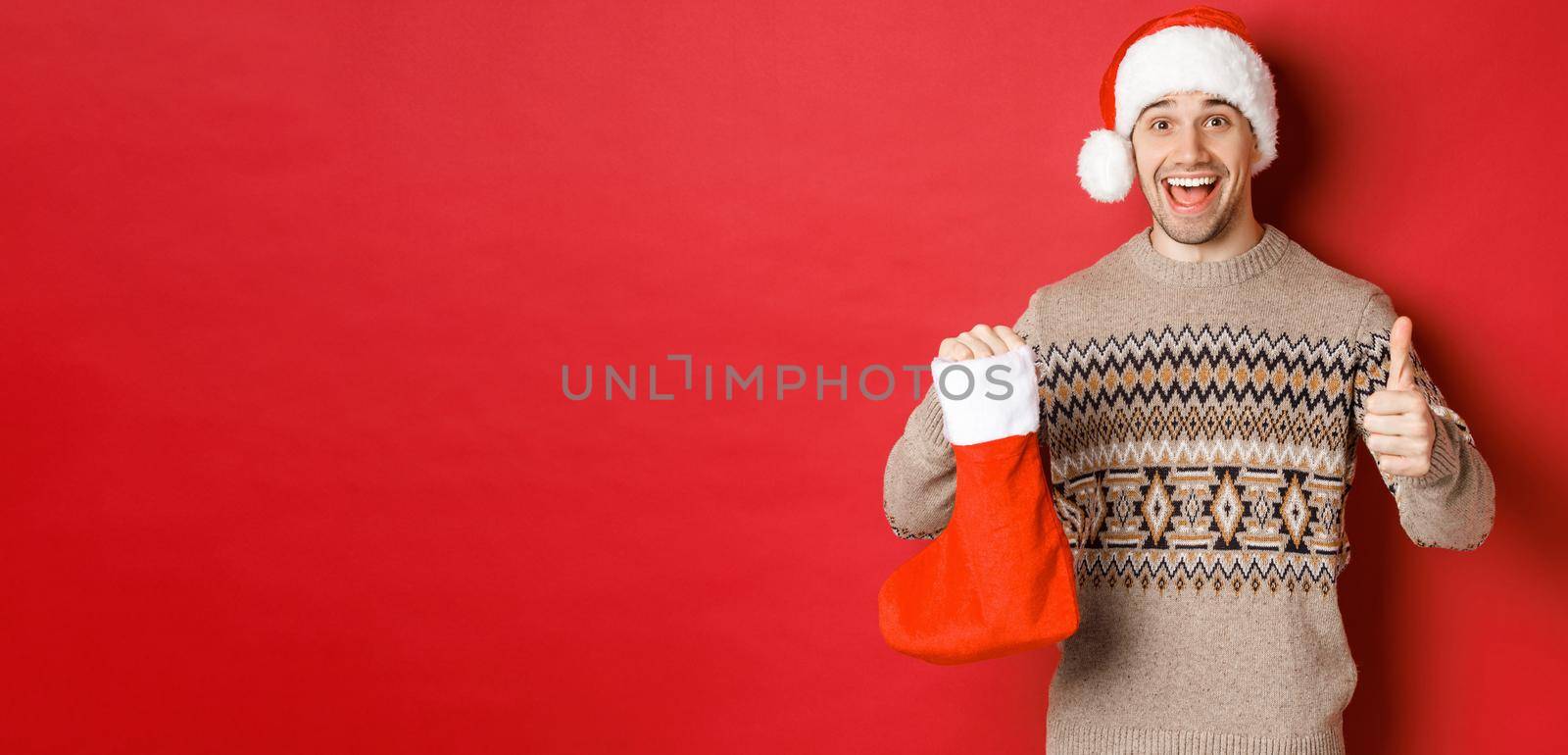 Concept of winter holidays, new year and celebration. Cheerful handsome man in santa hat and sweater, showing christmas stocking with candies and gifts, making thumbs-up.
