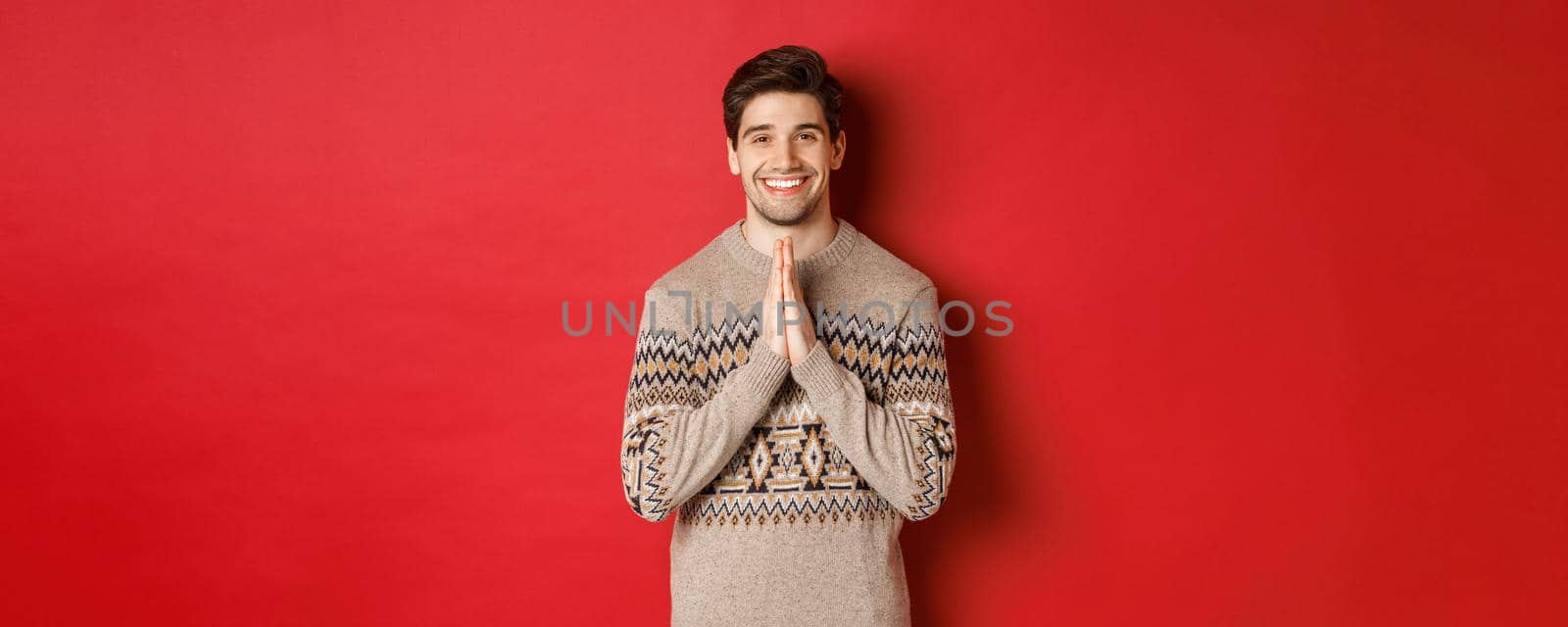 Image of happy and cute man in christmas sweater, asking for favour, holding hands together, smiling and saying thank you, feeling grateful, standing over red background by Benzoix