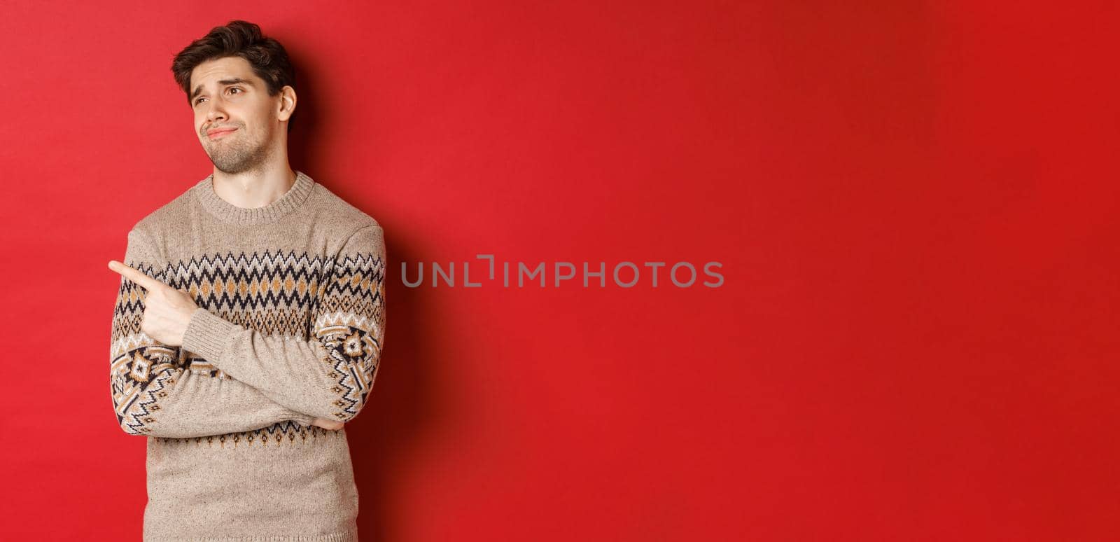 Image of disappointed handsome man in christmas sweater, looking and pointing left with upset face, express regret, showing new year advertisement, standing over red background by Benzoix