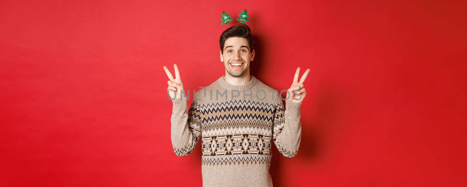 Concept of winter holidays, christmas and celebration. Image of handsome and silly guy dressed for new year party, showing peace signs and smiling, standing against red background by Benzoix