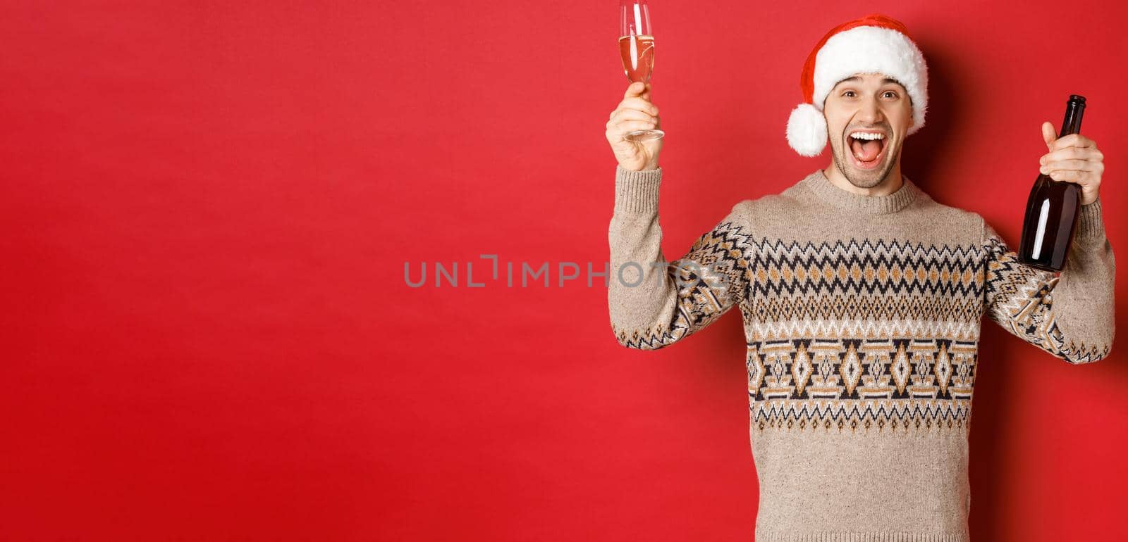 Concept of winter holidays, christmas and celebration. Excited handsome guy in sweater and santa hat, enjoying new year party, raising glass and champagne bottle, having fun.