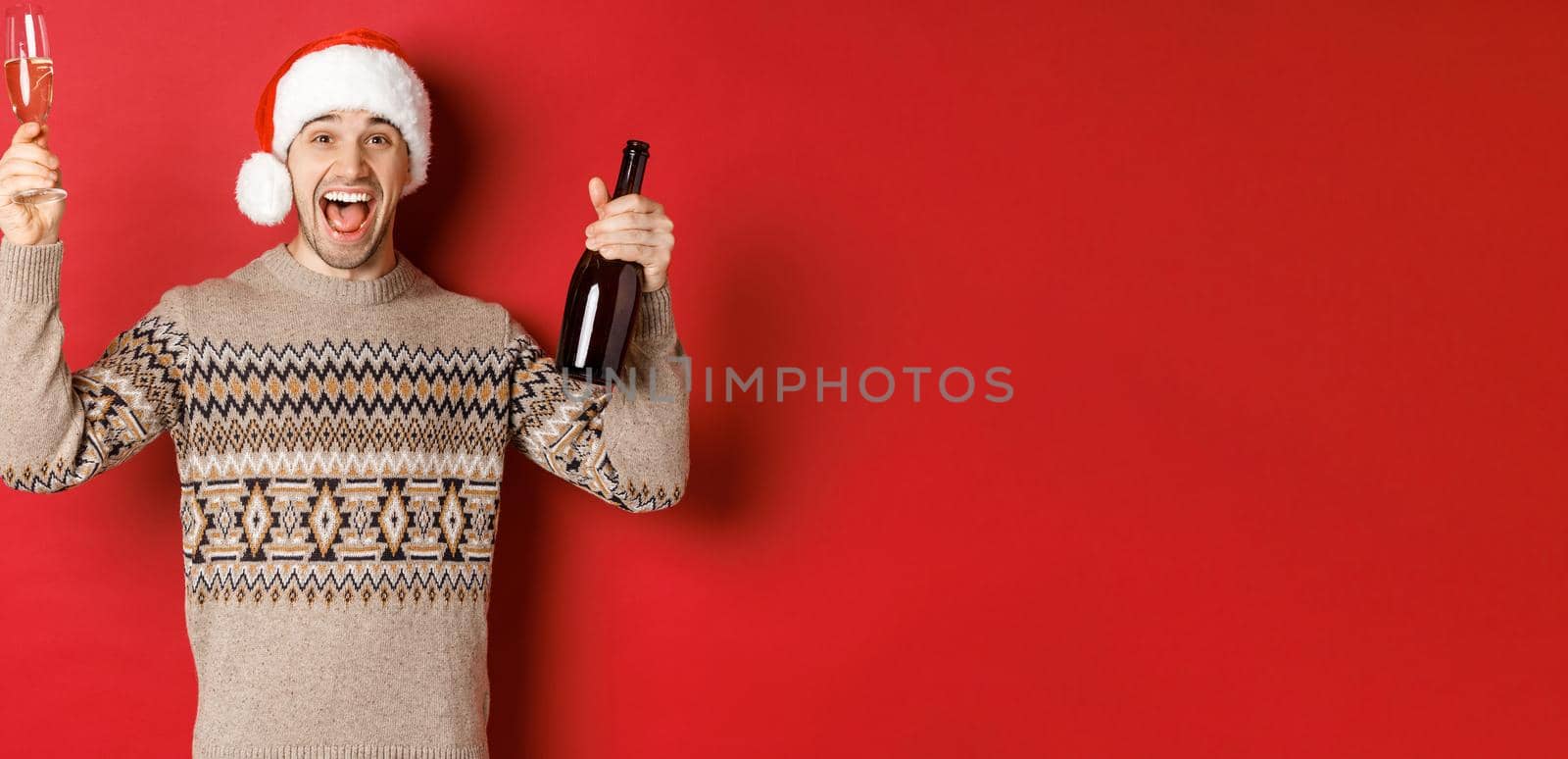 Concept of winter holidays, christmas and celebration. Excited handsome guy in sweater and santa hat, enjoying new year party, raising glass and champagne bottle, having fun.