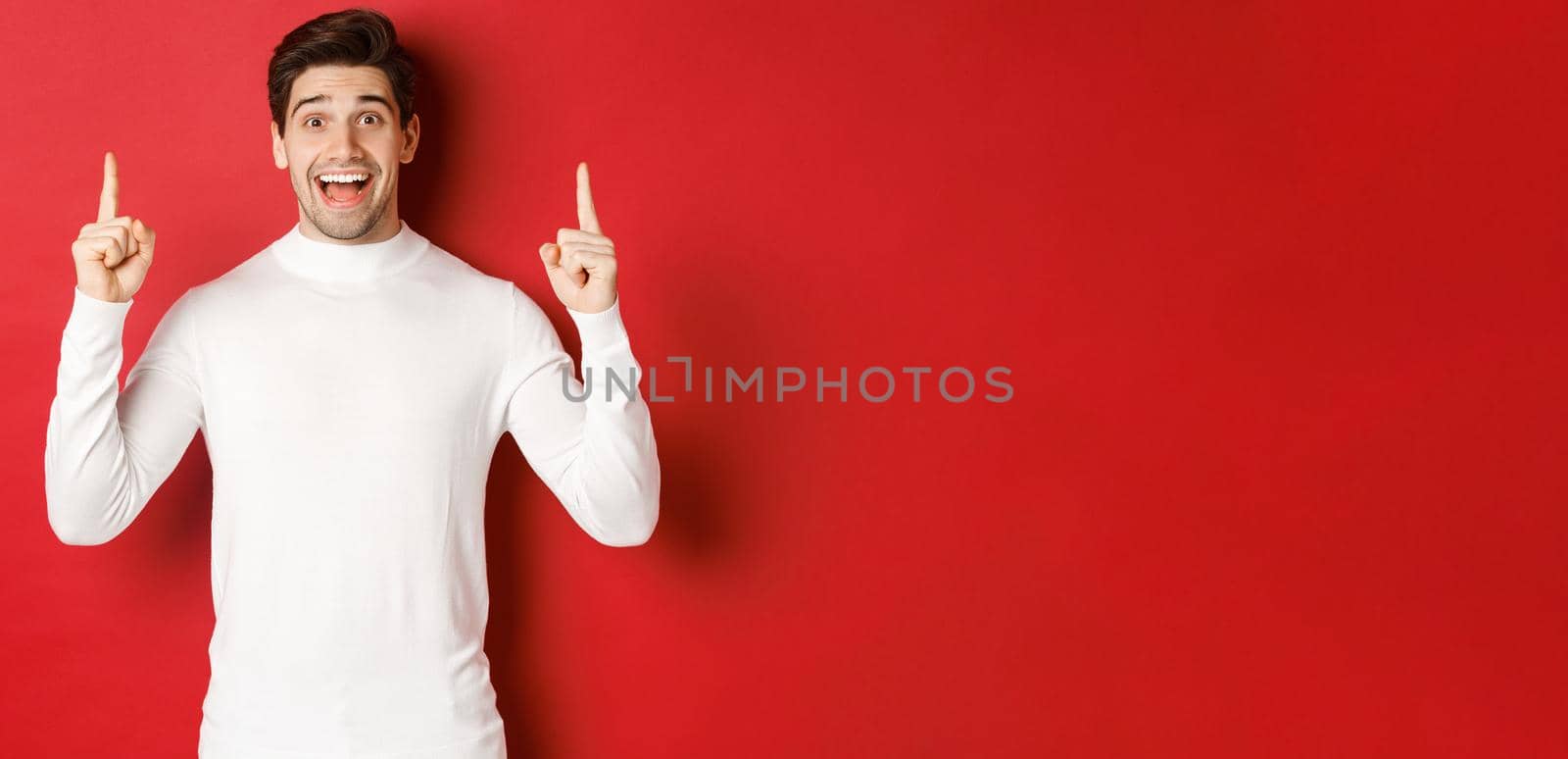Concept of winter holidays. Good-looking young man with bristle, wearing white sweater, showing christmas advertisement on copy space, pointing fingers up and looking amazed, red background by Benzoix
