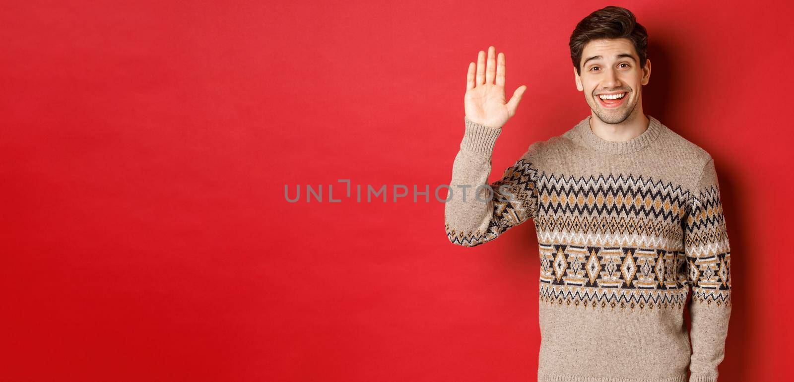 Portrait of handsome and friendly guy in christmas sweater, saying hello and waving hand, greeting someone and wishing happy winter holidays, standing over red background by Benzoix