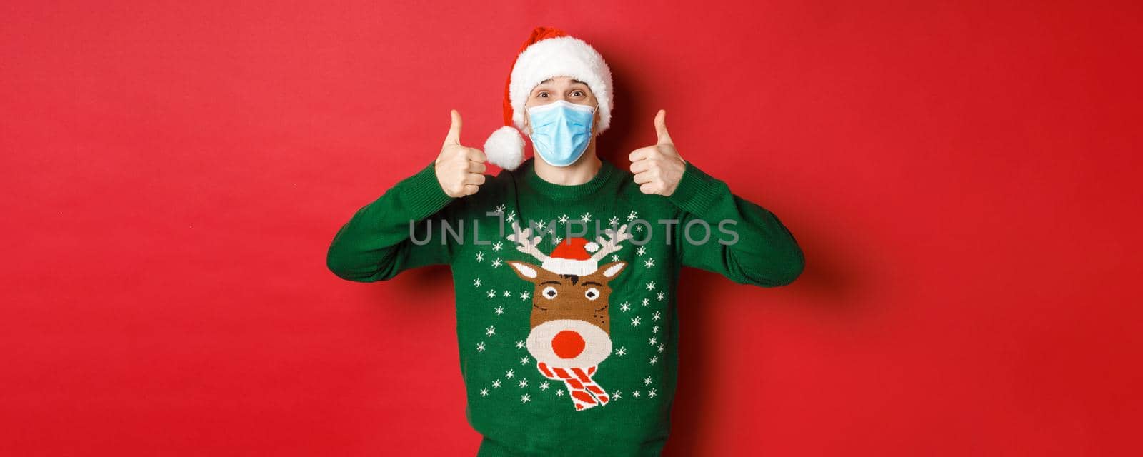 Concept of new year, covid-19 and social distancing. Cheerful handsome man in santa hat and medical mask, showing thumbs-up, enjoying christmas party, standing over red background by Benzoix