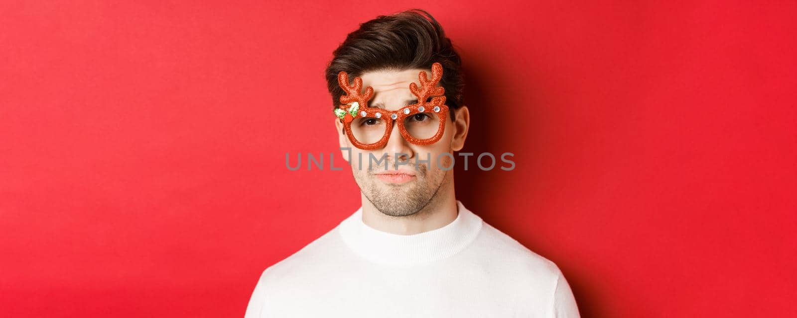 Concept of winter holidays, christmas and celebration. Close-up of skeptical handsome guy, wearing party glasses, smirk and looking unamused, standing over red background.