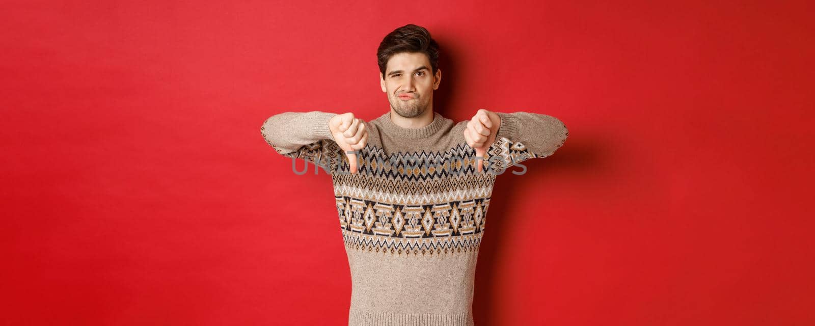 Portrait of skeptical and disappointed handsome man in christmas sweater, dislike new year party, showing thumbs-down, express disapproval, standing over red background by Benzoix