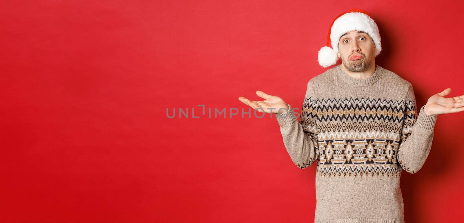 Image of clueless young man in sweater and santa hat, shrugging and looking unaware, dont know what to buy for new year holidays, standing over red background.