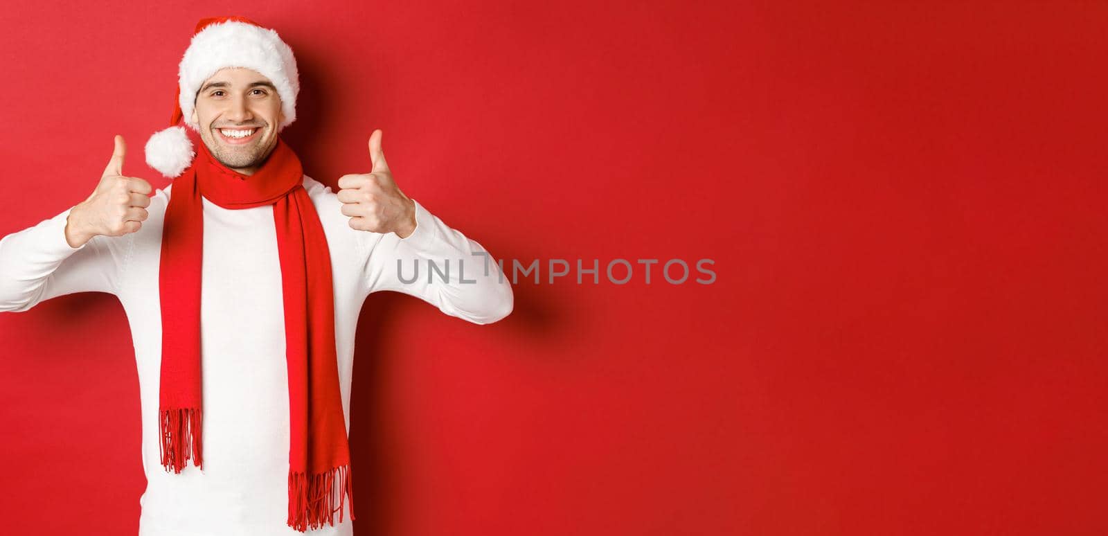 Portrait of handsome smiling man in santa scarf and hat, showing thumbs-up, celebrating christmas, standing over red background.