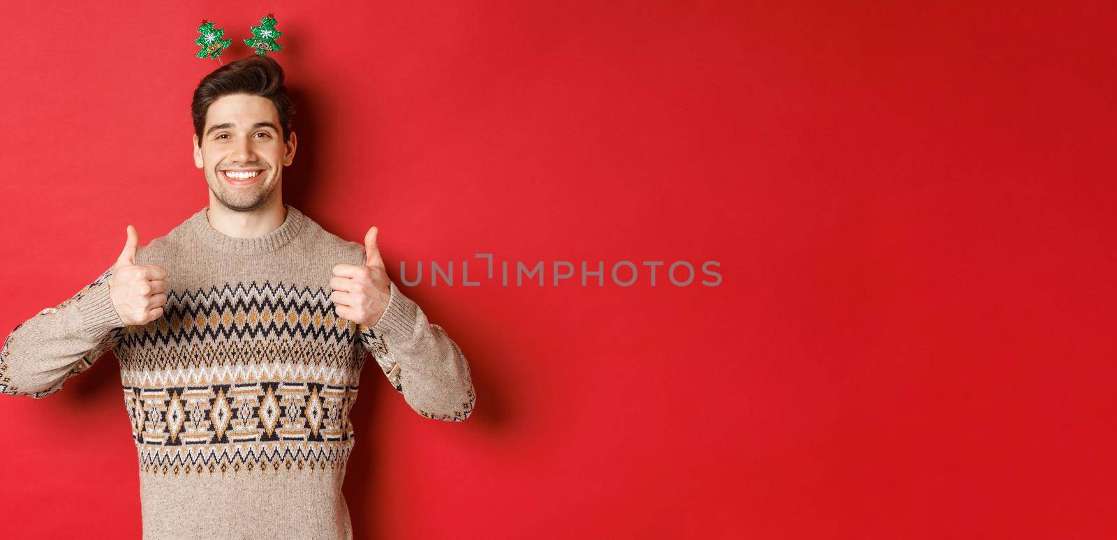 Concept of winter holidays, christmas and celebration. Cheerful bearded guy in sweater, showing thumbs-up in approval and smiling, enjoying new year party, red background by Benzoix