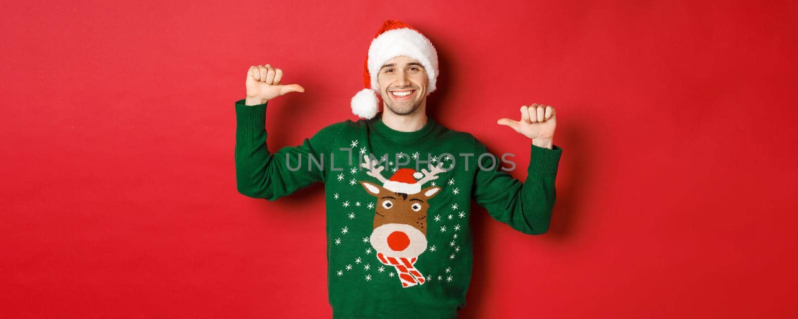 Concept of winter holidays, christmas and lifestyle. Happy attractive guy in santa hat and sweater, pointing at himself with pleased face, standing over red background.