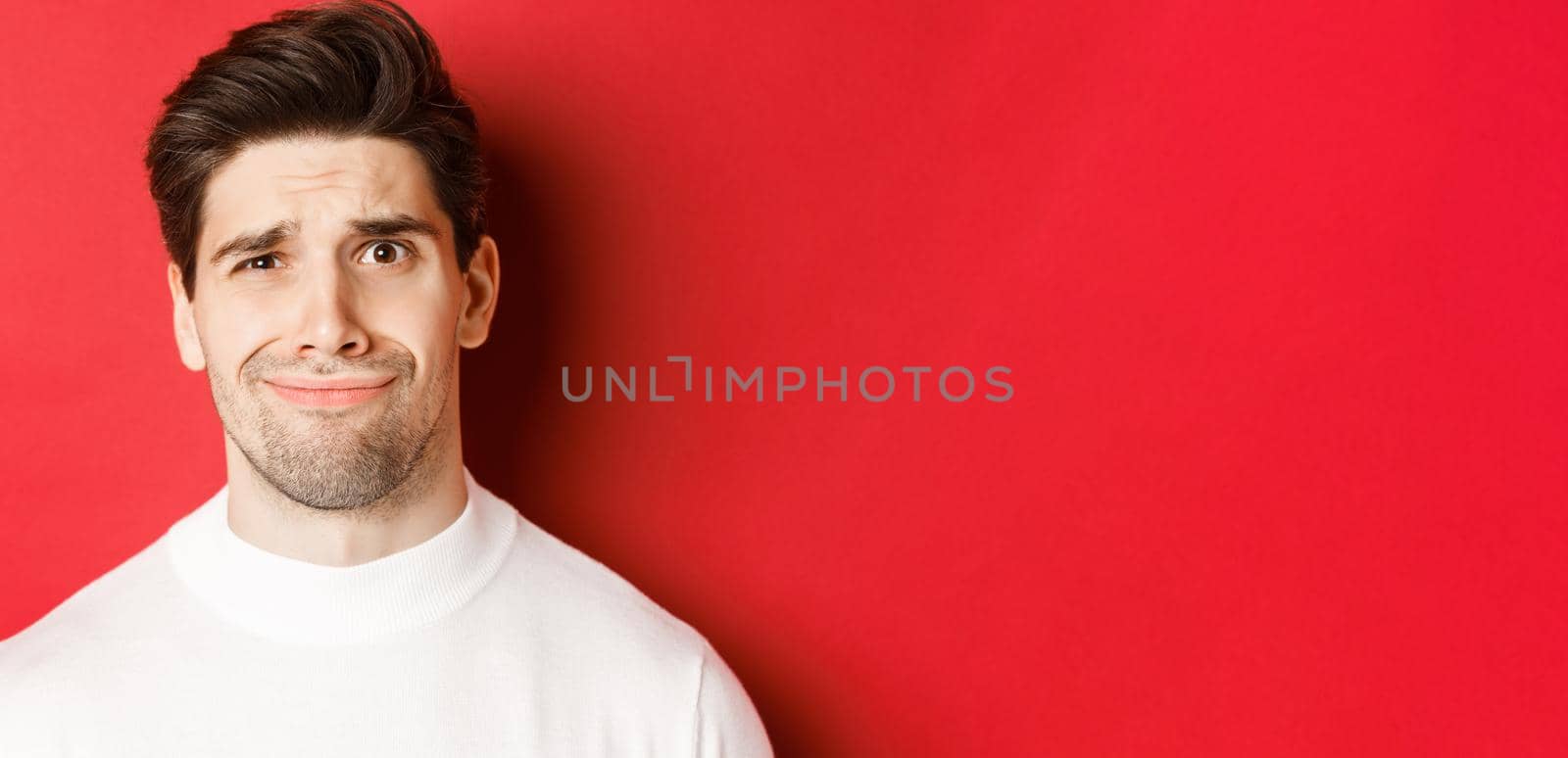 Close-up of handsome man feeling uncomfortable, grimacing and looking at something displeasing, standing over red background by Benzoix