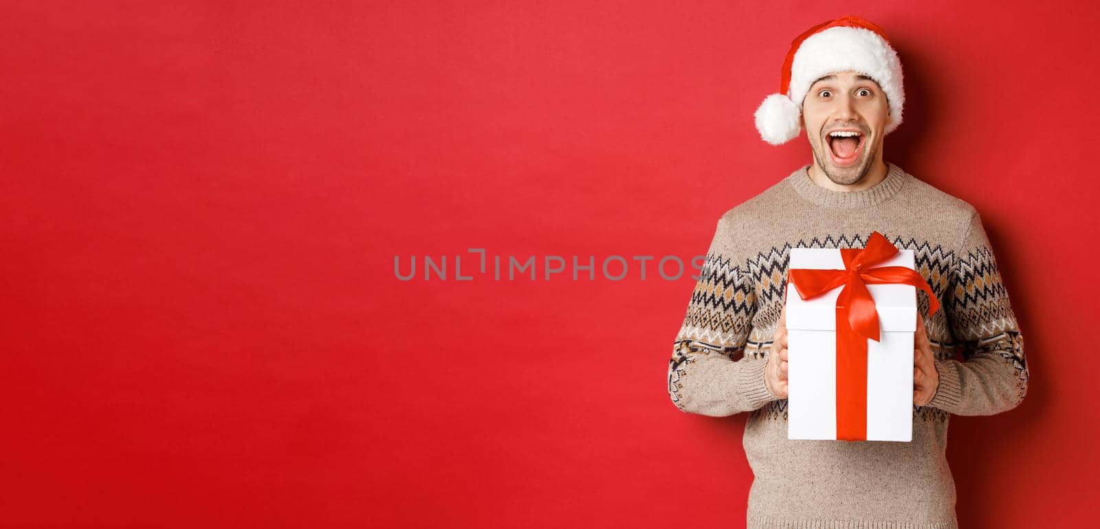 Image of excited handsome man receiving christmas gift, wearing santa hat and winter sweater, shouting for joy, holding present and standing over red background by Benzoix