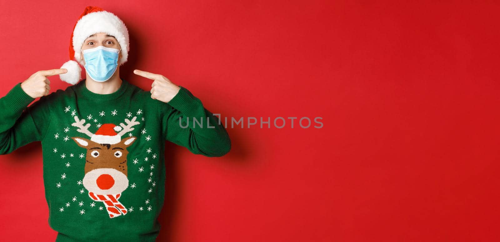 Concept of new year, coronavirus and social distancing. Happy man in santa hat and christmas sweater, recommending to wear medical mask on party, standing over red background by Benzoix