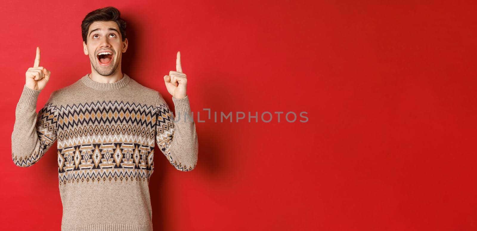 Image of excited handsome guy who likes winter holidays showing advertisement, looking and pointing fingers up with amazed face, standing in christmas sweater over red background.