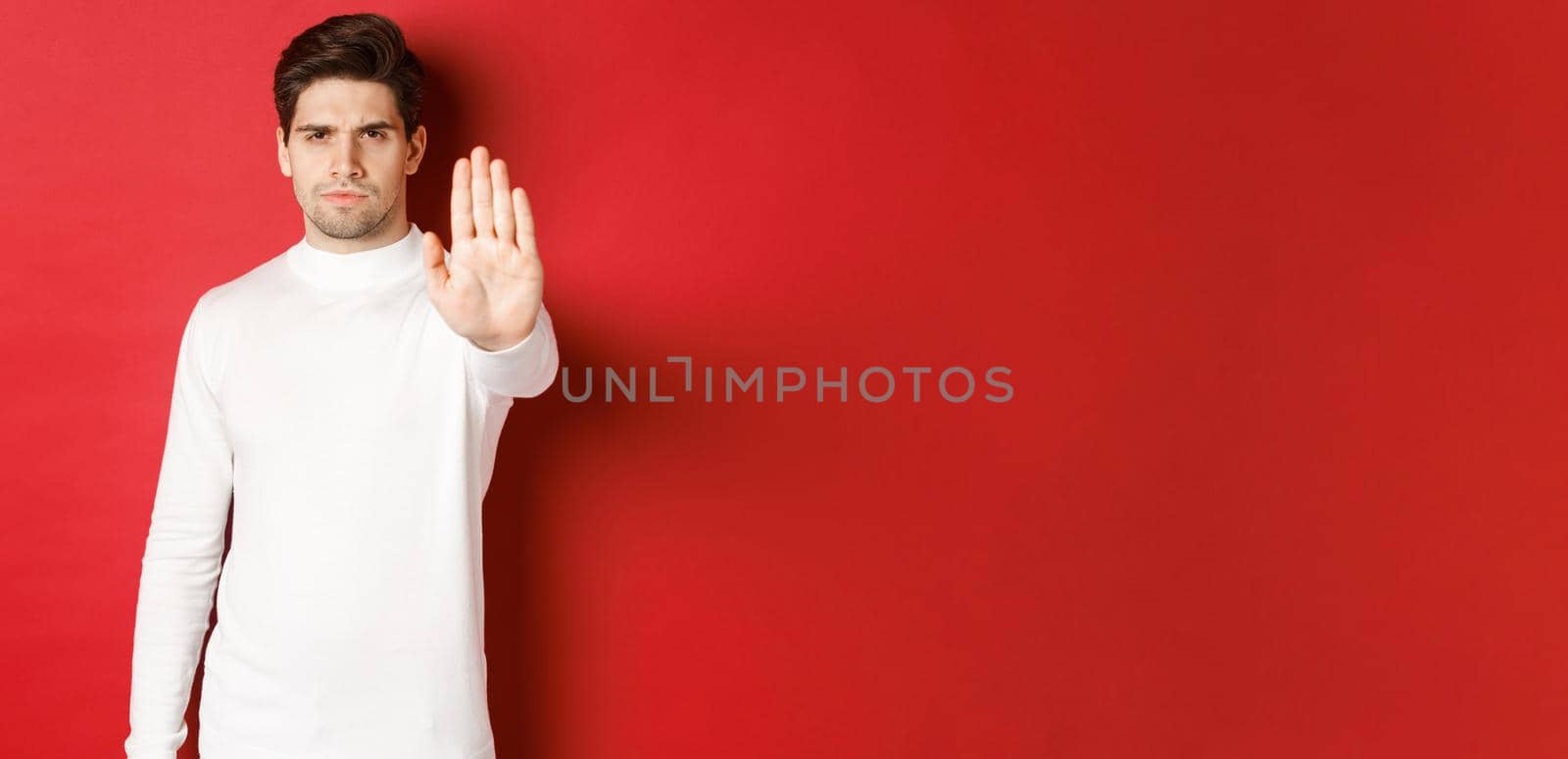 Image of serious and confident man telling to stop, forbid something, extending one hand and prohibit action, standing over red background.