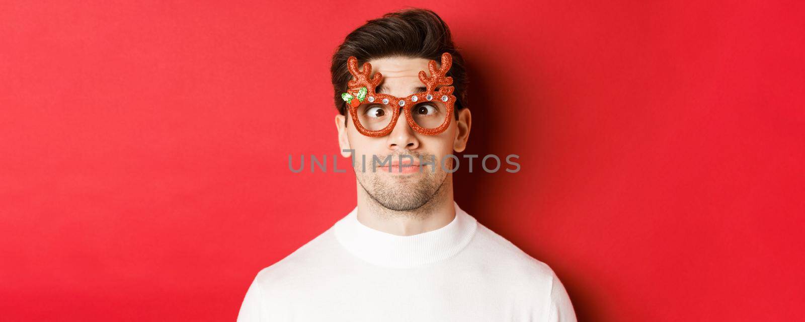 Concept of winter holidays, christmas and celebration. Close-up of funny brunette in party glasses, squinting and making faces, standing over red background.