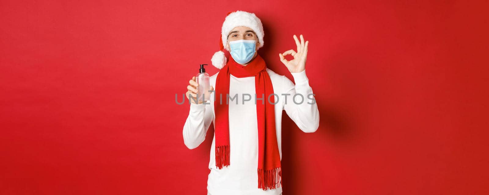 Concept of covid-19, christmas and holidays during pandemic. Attractive man in santa hat and medical mask, showing okay sign while recommending hand sanitizer, standing over red background.