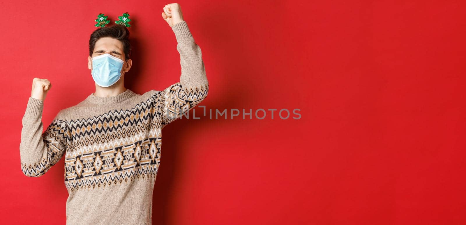 Concept of coronavirus, winter holidays and social distancing. Image of excited and happy man in medical mask and christmas clothing, raising hands up and celebrating new year, red background.