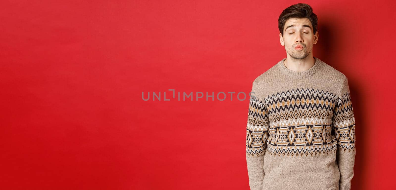 Image of handsome man in christmas sweater pucker lips and close eyes, waiting for kiss under mistletoe, standing over red background.