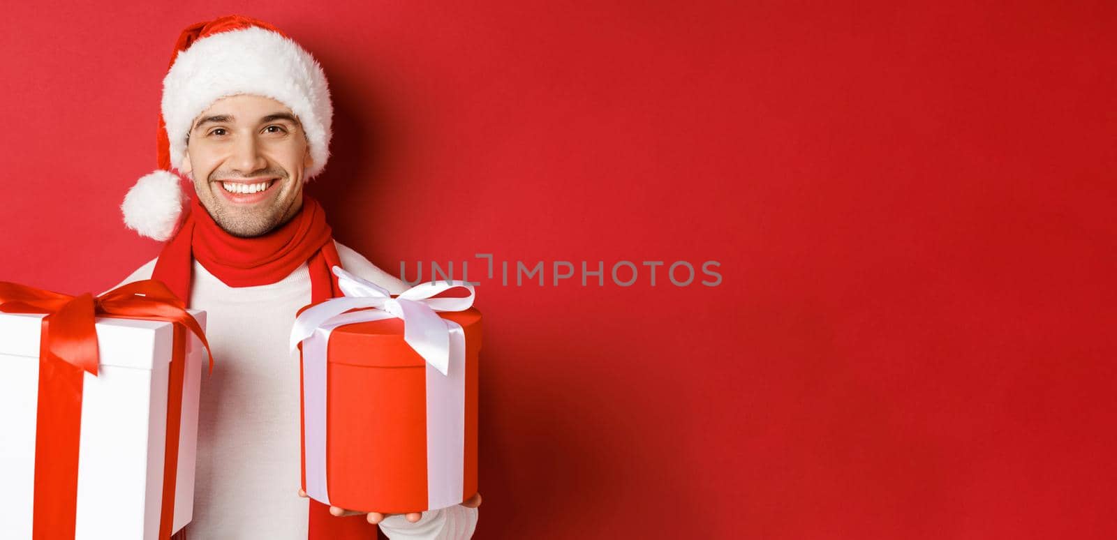 Concept of winter holidays, christmas and lifestyle. Close-up of smiling handsome guy in santa hat and scarf, wishing happy new year and holding gifts, standing with presents over red background by Benzoix